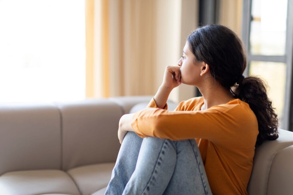 Woman sitting on a couch with her knees to her chest, chin on her hand, looking out the window.