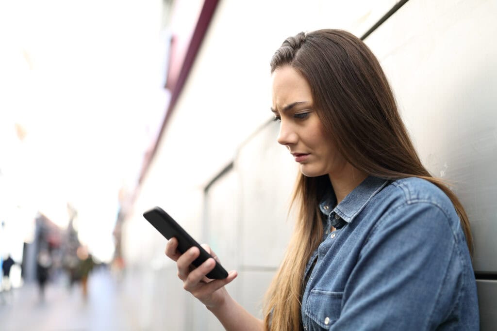 Woman with long brown hair standing against building looking at her smart phone looking confused.