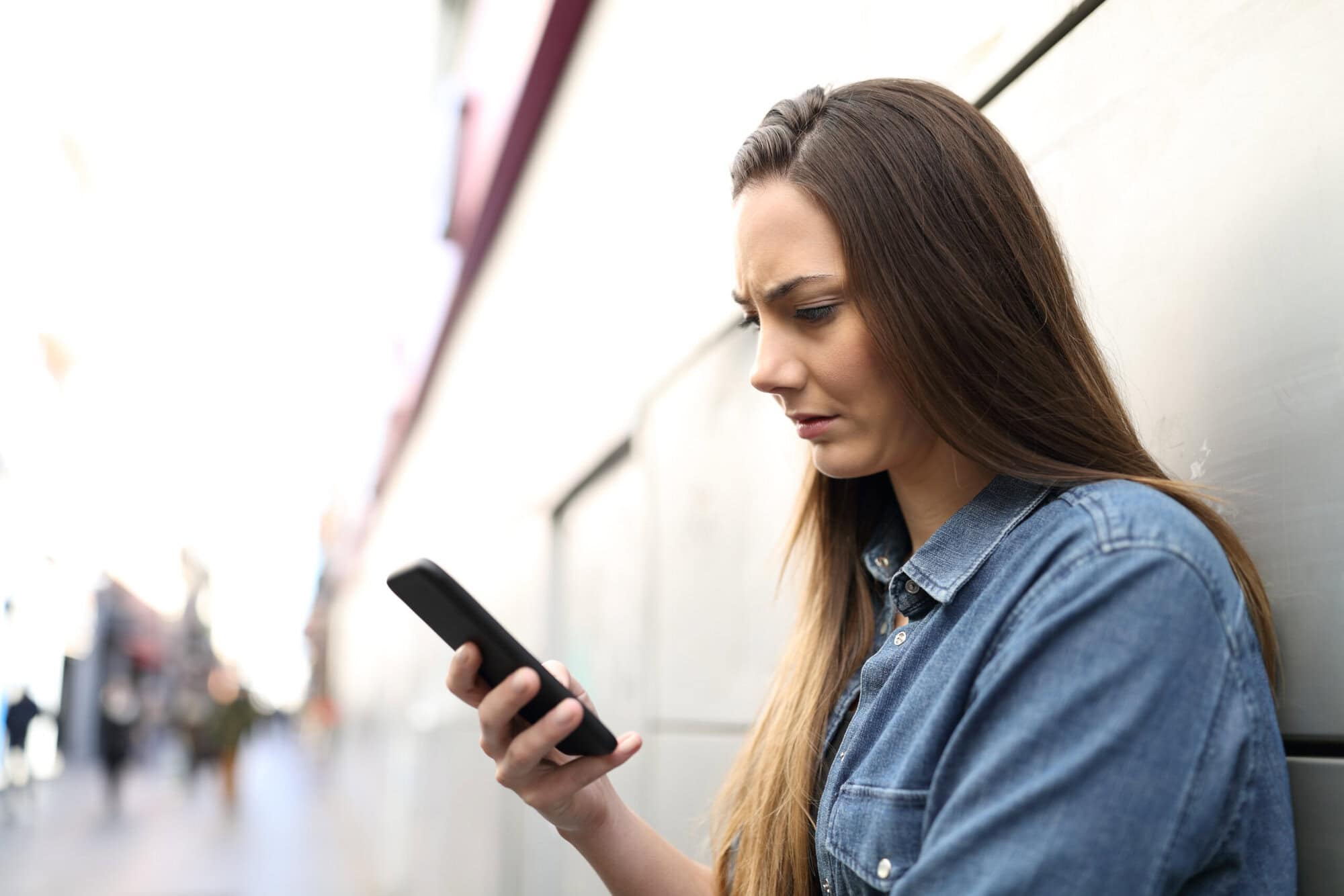 Woman with long brown hair standing against building looking at her smart phone looking confused.
