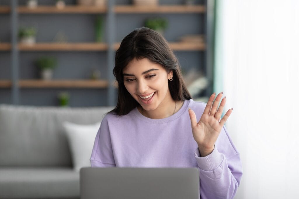 Young woman with brown hair and a purple sweatshirt waving to laptop computer, talking to therapist, with a smile.