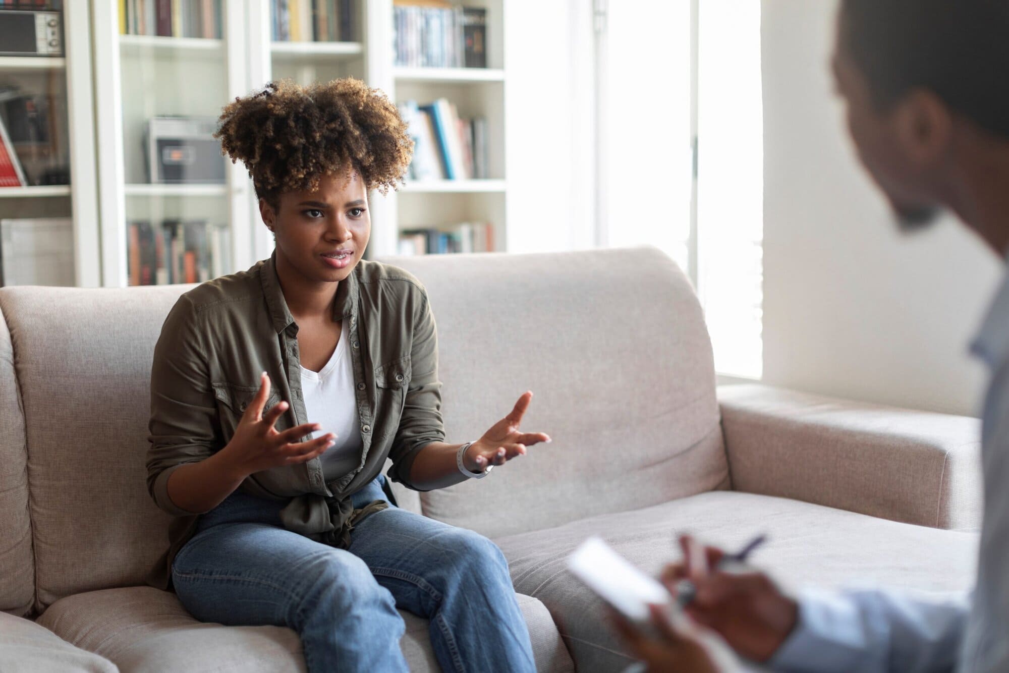Woman sitting on the couch talking to a therapist. The therapists hands holding a pen and paper are visible.
