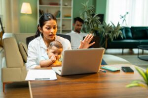Woman sitting at table talking to person on a computer while holding her baby. Her husband is on the couch in the background.