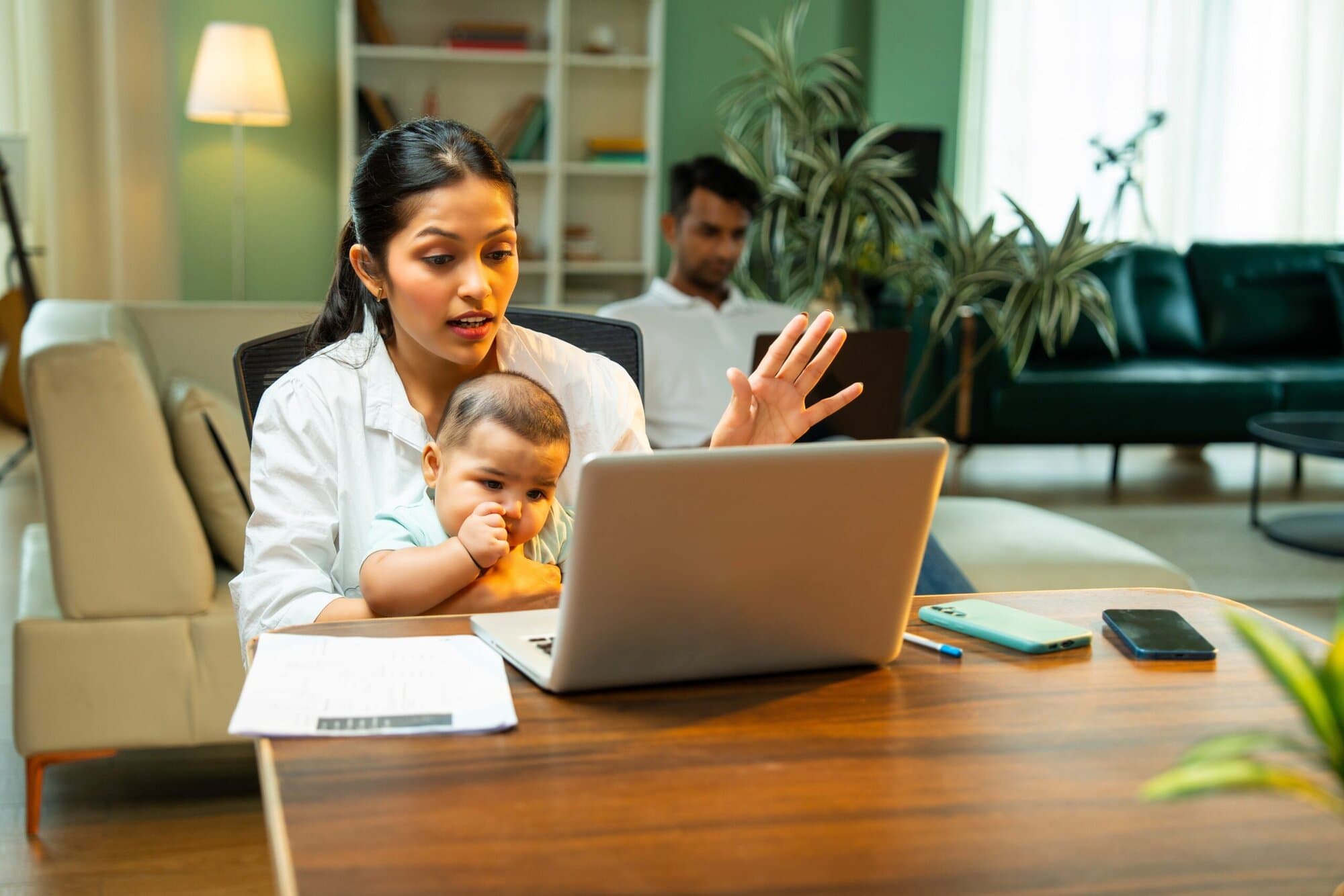 Woman sitting at table talking to person on a computer while holding her baby. Her husband is on the couch in the background.