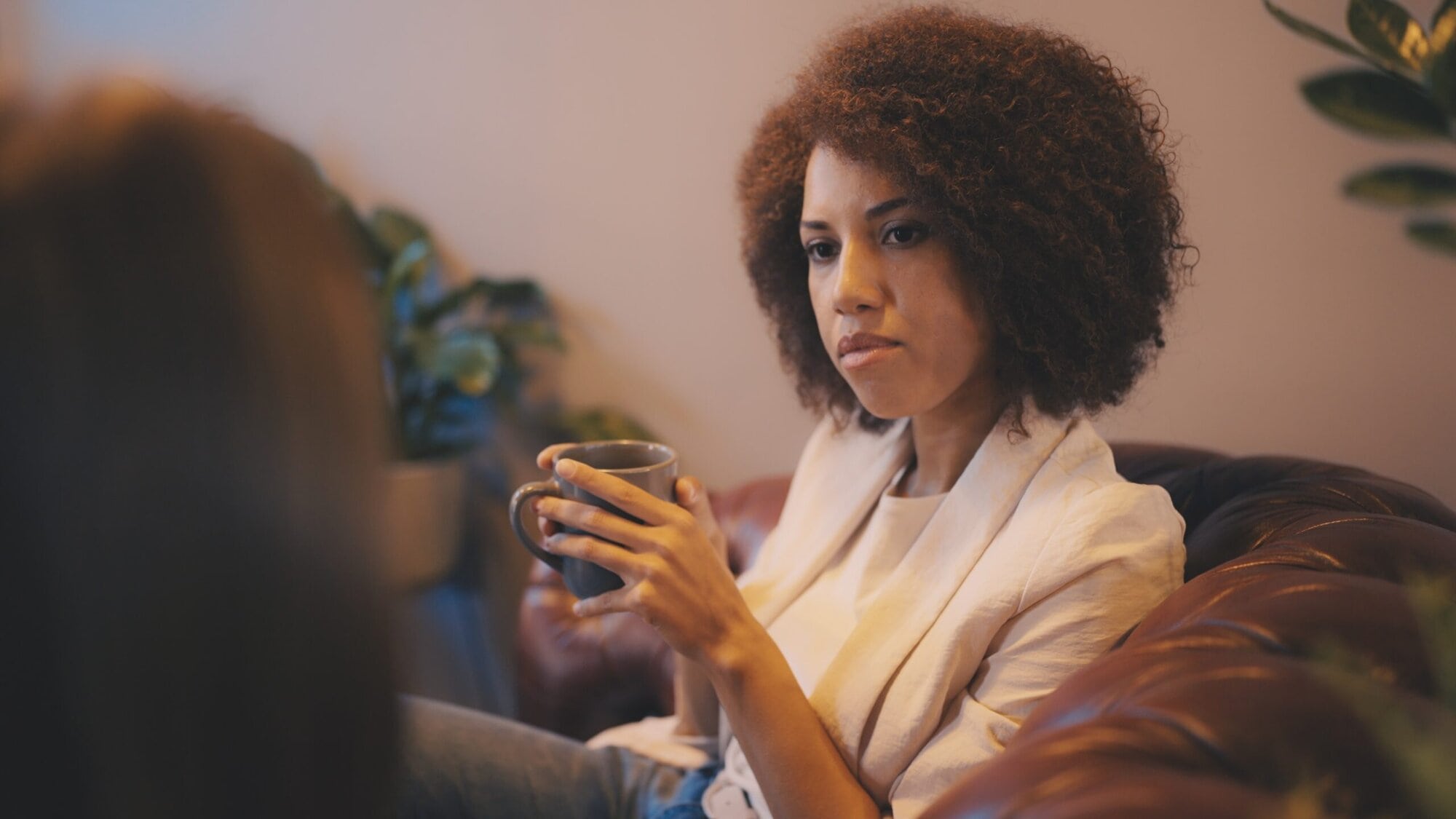 Woman sitting on the couch holding a mug talking to someone out of camera frame.
