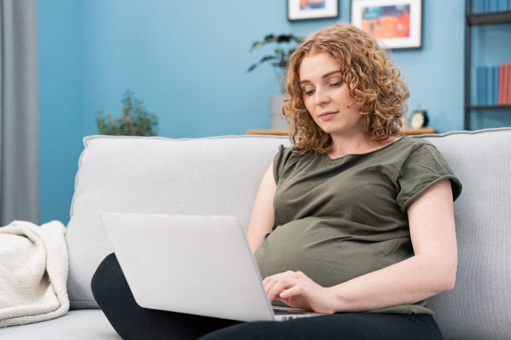 Pregnant woman with curly brown hair sitting on couch looking at laptop.