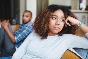 Woman sitting on couch with man behind her. He is looking at her, she is looking away. Her hand is in her head.