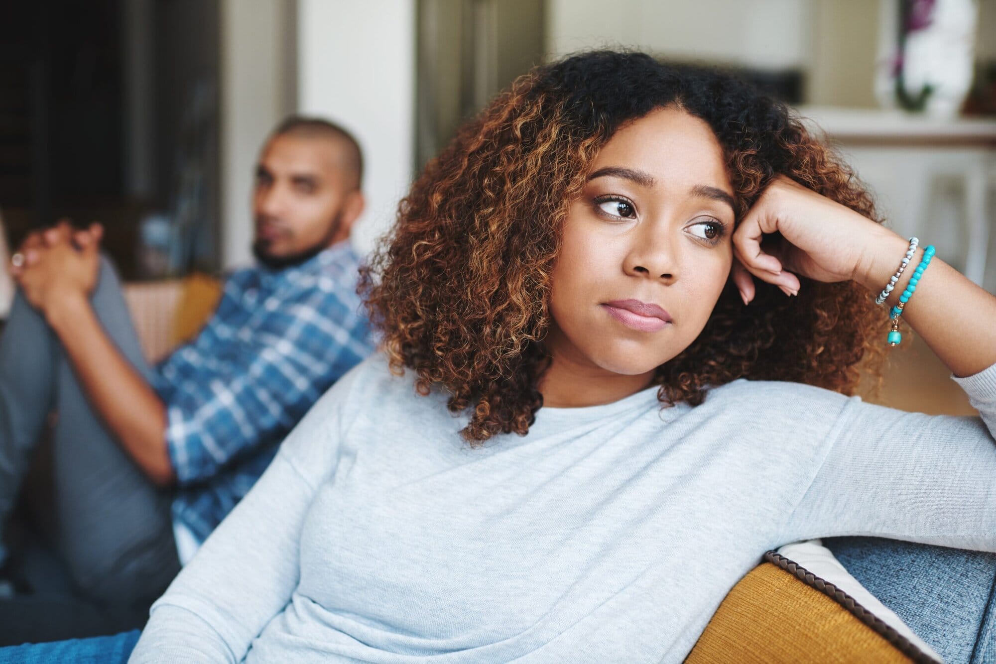 Woman sitting on couch with man behind her. He is looking at her, she is looking away. Her hand is in her head.