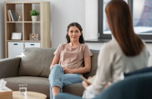 Two women sitting looking at each other. One has back to camera. One on a chair, one on a couch.
