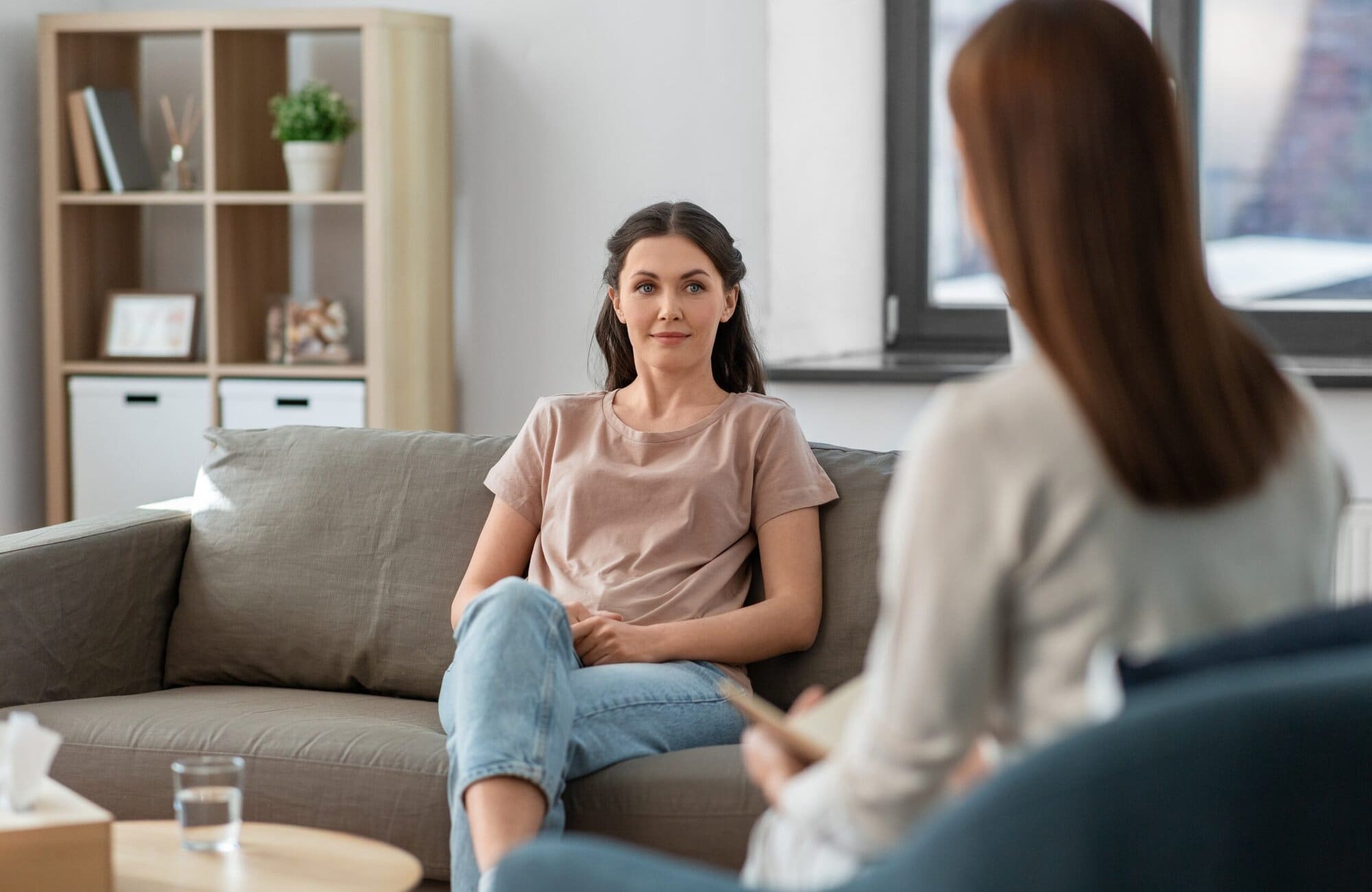 Two women sitting looking at each other. One has back to camera. One on a chair, one on a couch.