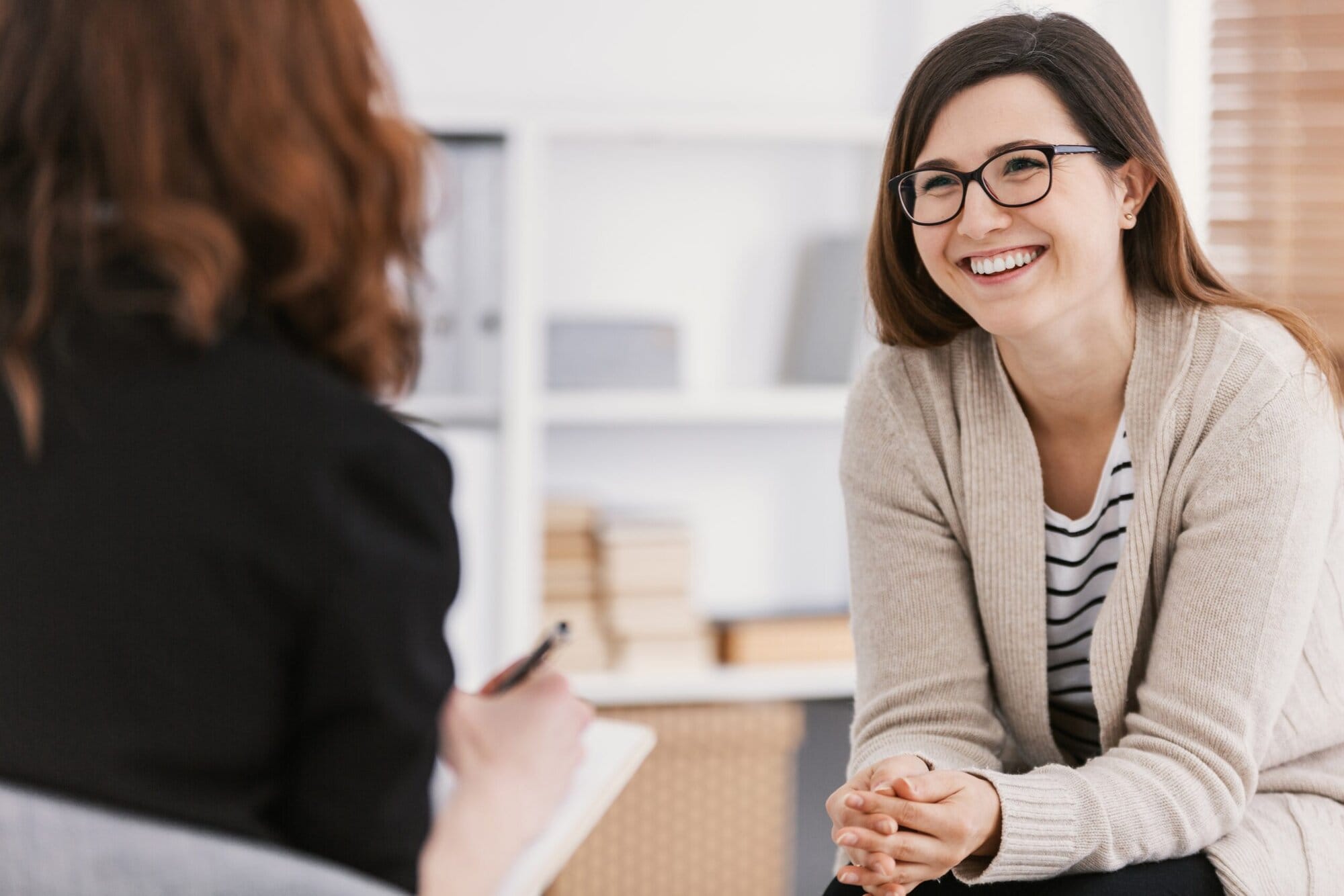 Two women sitting in chairs looking at each other. One with back to camera has notebook and pen in her hands. The other is smiling..