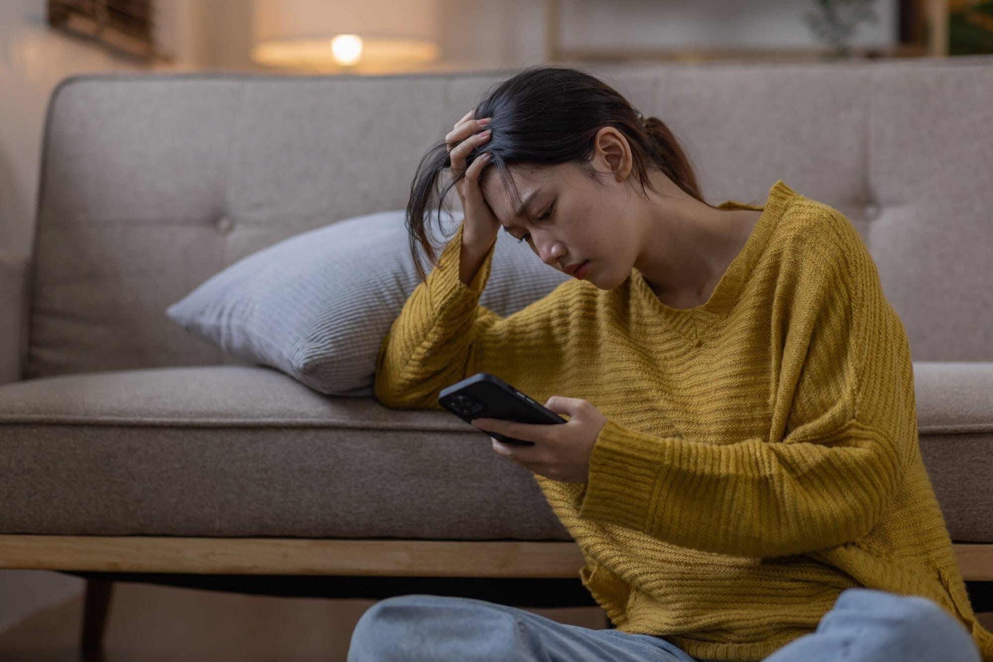 Woman sitting on the floor with her right elbow on the couch and right hand in her head. She is looking at her phone in her left hand.