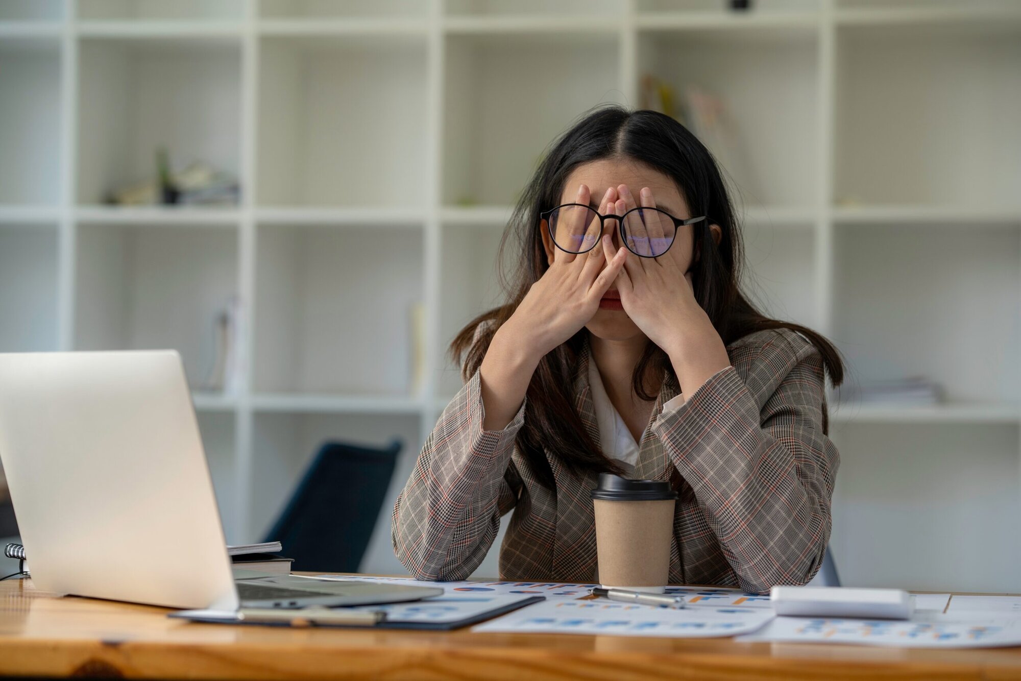 Woman sitting at her desk in front of computer with coffee. Head in her hands under her eye glasses, rubbing her eyes.