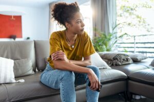 Woman sitting on a couch leaned forward slightly. Left arm on right arm and looking out the window to the left of her.