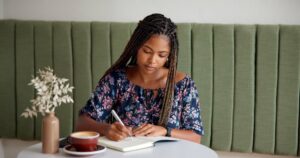 Woman sitting at table writing in a notebook with coffee cup next to her.
