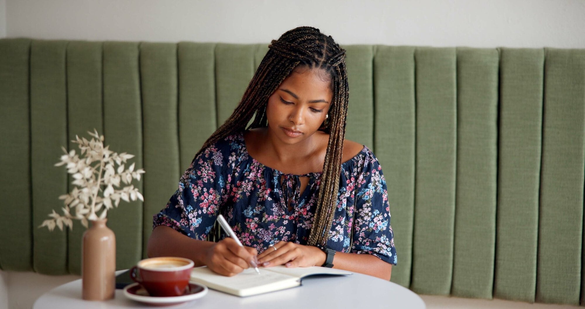 Woman sitting at table writing in a notebook with coffee cup next to her.