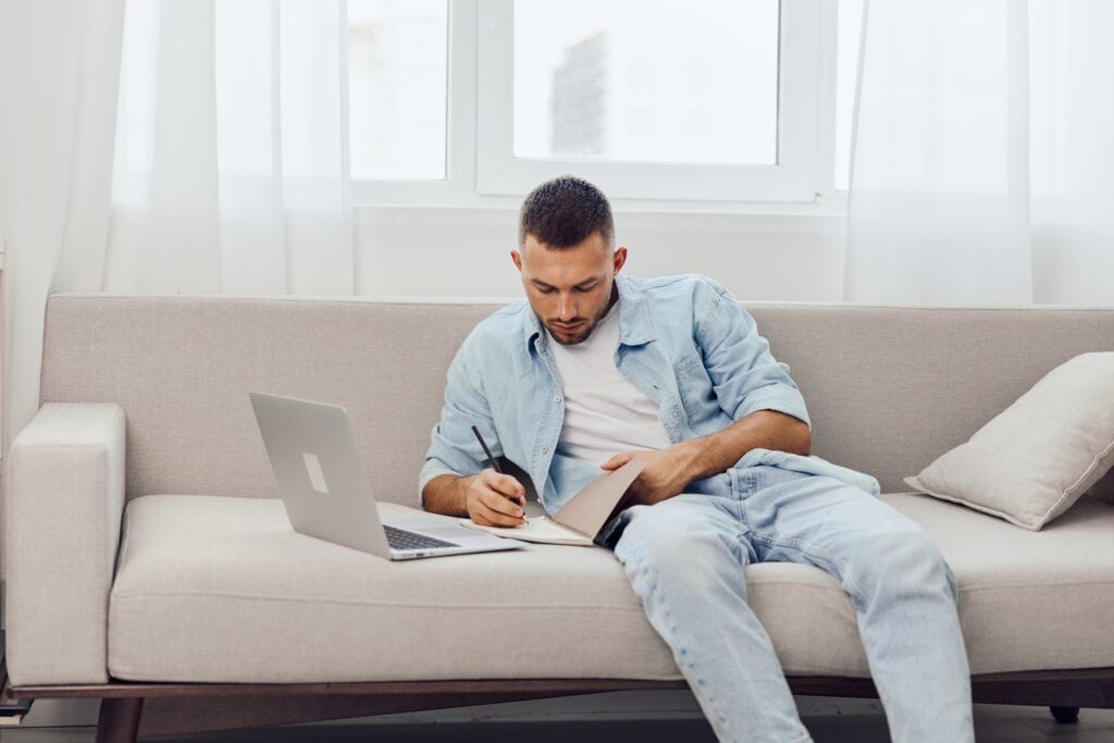 Man sitting on couch writing in notebook with laptop next to him.