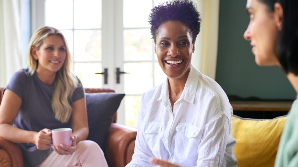 Three women sitting on couches and chairs looking at eachother. 2 are smiling, 1 is holding coffee.
