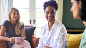 Three women sitting on couches and chairs looking at eachother. 2 are smiling, 1 is holding coffee.