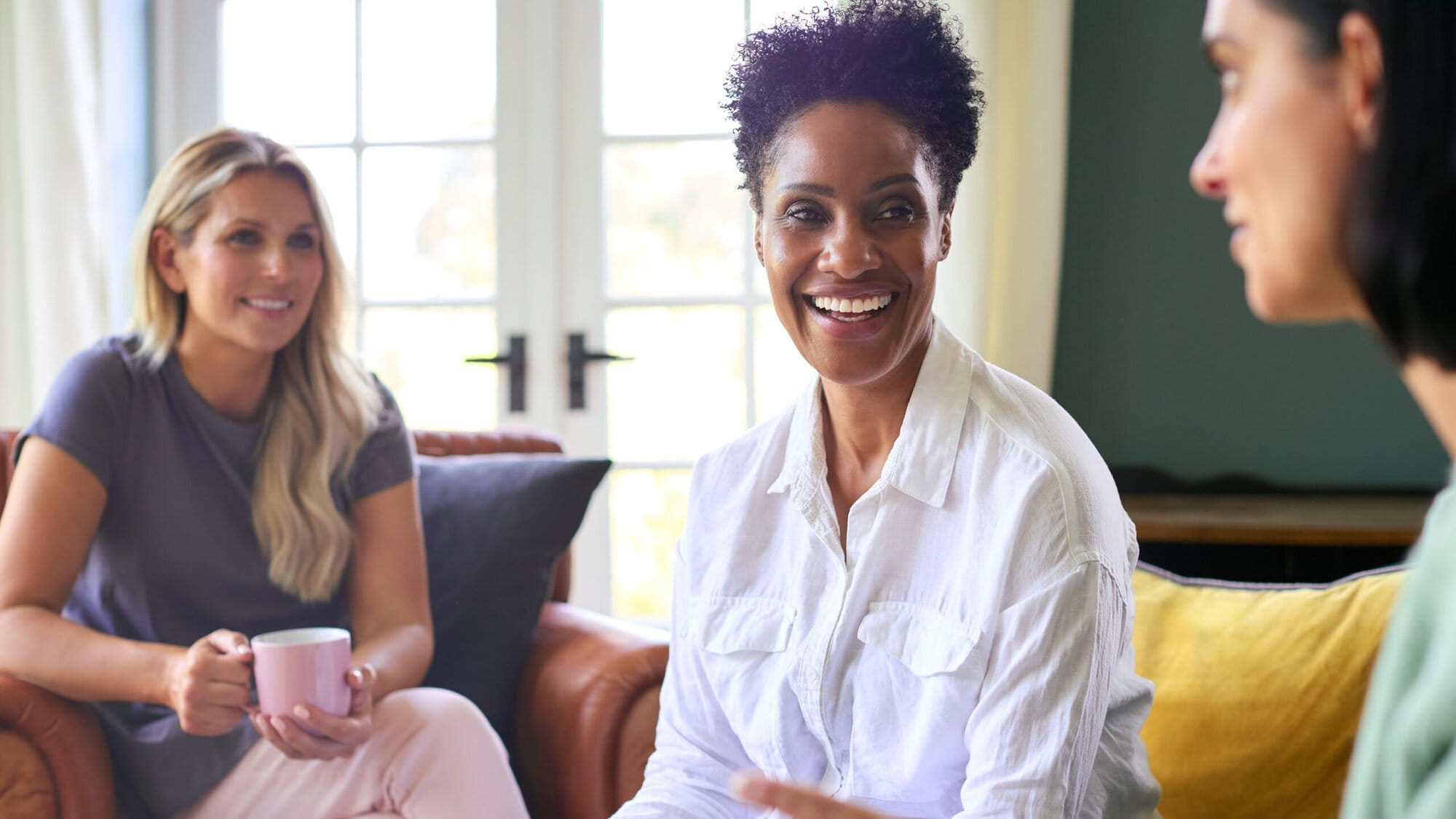 Three women sitting on couches and chairs looking at eachother. 2 are smiling, 1 is holding coffee.