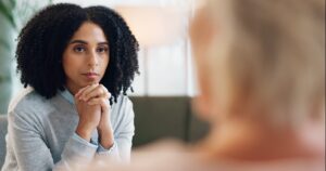 Woman sitting looking at another women whose back of head is to the camera. Woman looking at her with hands folded under chin.