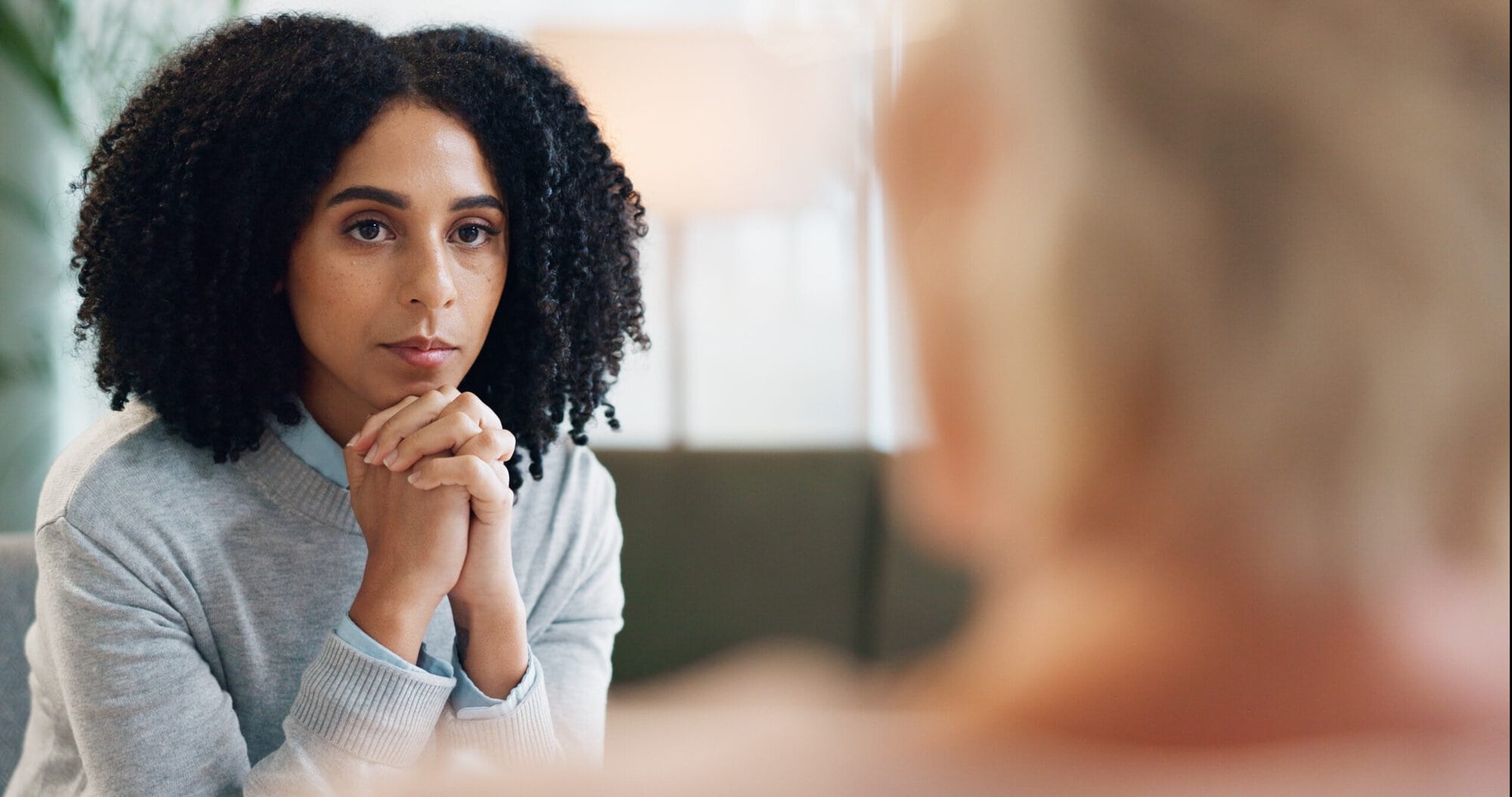 Woman sitting looking at another women whose back of head is to the camera. Woman looking at her with hands folded under chin.